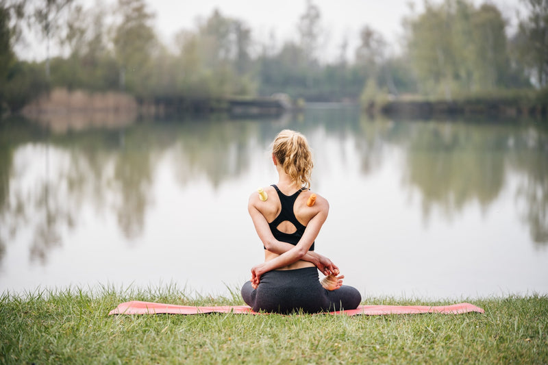 Eine Person sitzt in einer Yoga-Pose vor einem See. Am Rücken sind zwei BellaBambi Cups platziert